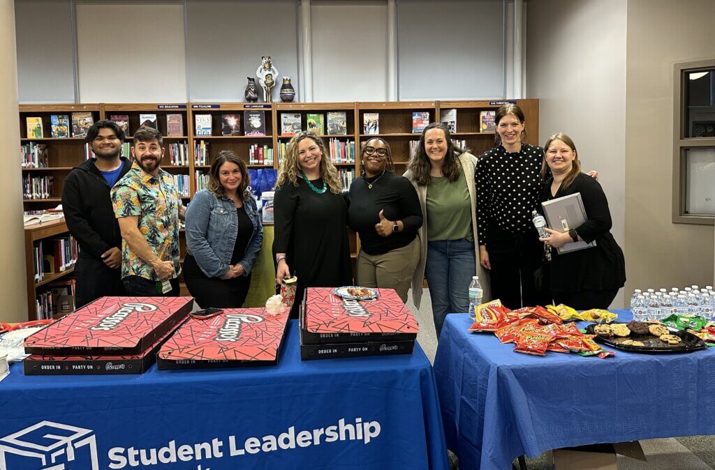 Eight staff members stand behind tables with pizza boxes, snacks, and drinks in a library, with a blue Student Leadership tablecloth in front.