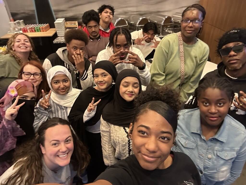A group of diverse people pose closely together indoors, smiling and making peace signs for a selfie, at the school's alumni day
