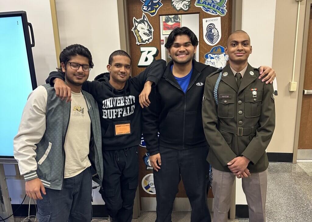 Four young men stand in front of a college counseling office, smiling at the camera; one wears a military uniform while the others are dressed casually.