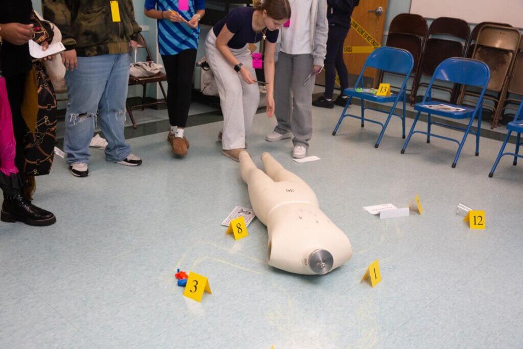 Students at at TYWLS Astoria stand around a mannequin on the floor of a classroom marked as a mock crime scene, with numbered evidence markers and chalk outline