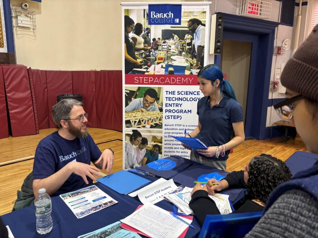 A man at a Baruch College STEP Academy table talks to two students at a summer enrichment fair