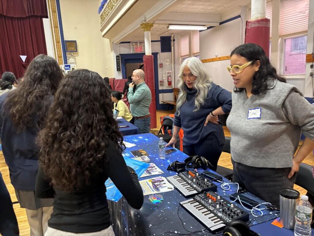 Two women stand behind a table with audio equipment, keyboards, and brochures, speaking to students at a summer enrichment fair