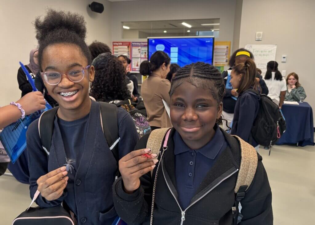 Two girls wearing backpacks stand indoors, smiling at the camera and holding up small objects. Other people and informational displays are visible in the background.