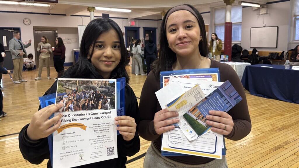 Two young women stand indoors holding informational flyers and pamphlets at an event with tables and other attendees in the background.