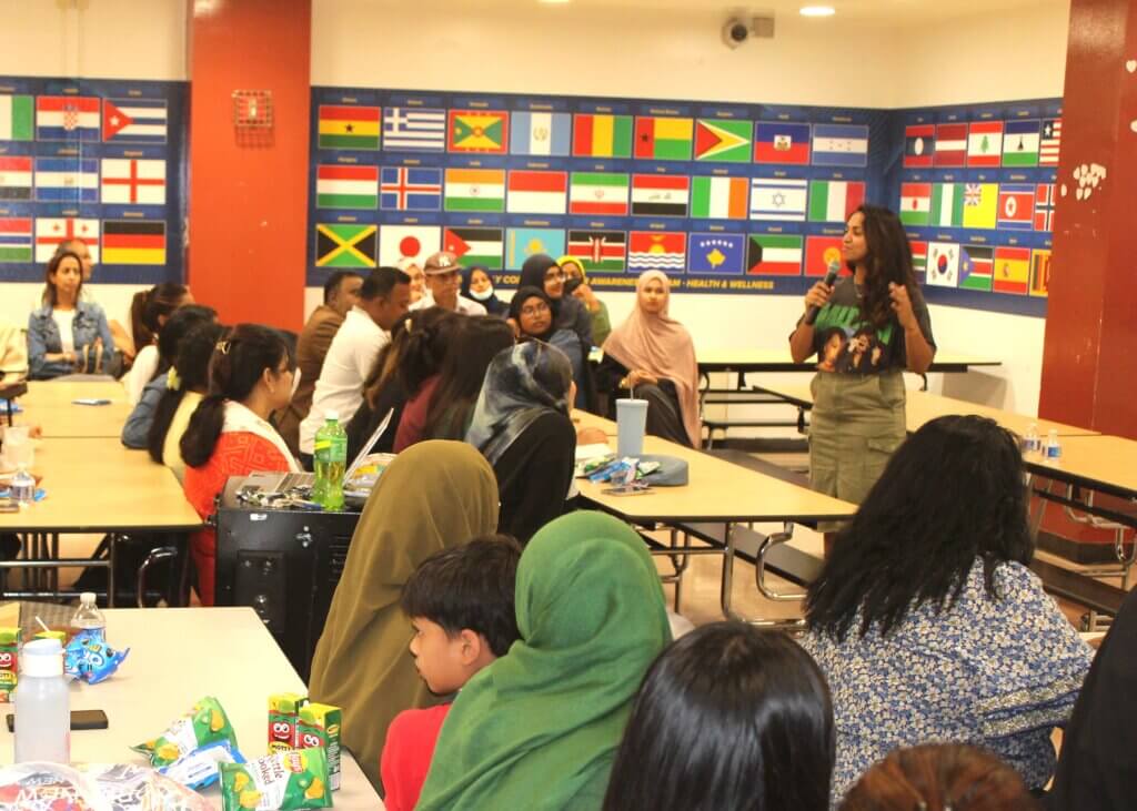 A college counselor speaks to a seated audience of families with children in a cafeteria decorated with international flags.