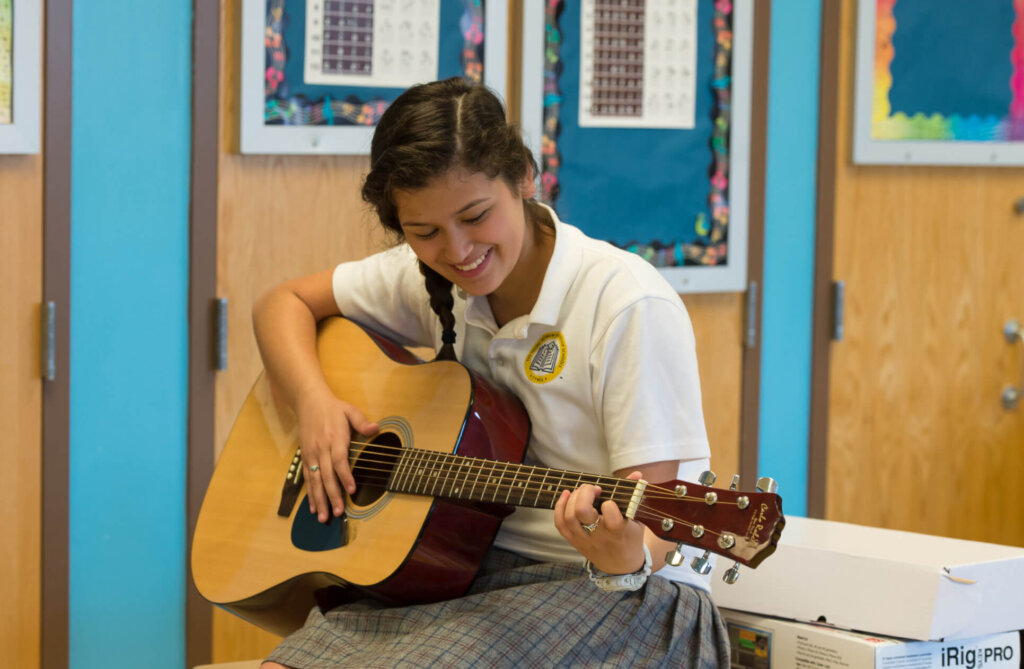 A student wearing a school uniform sits indoors and smiles while playing an acoustic guitar.