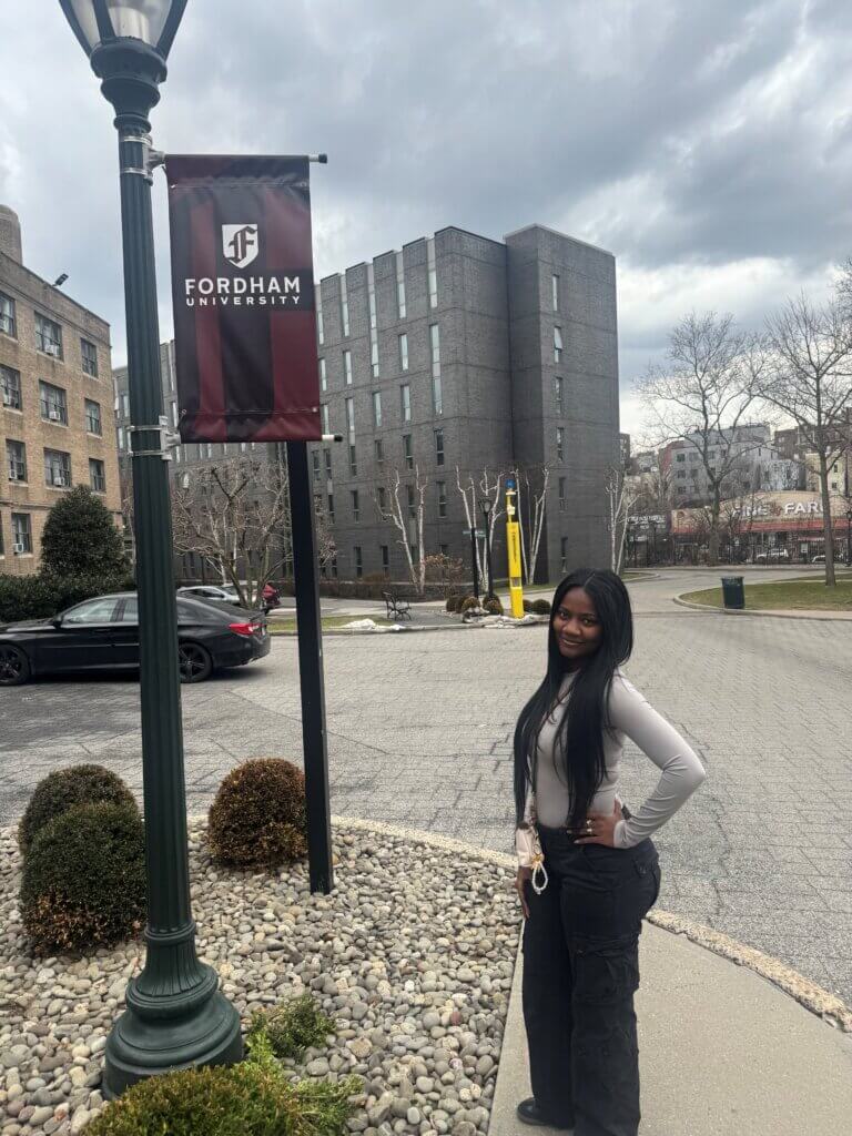 A woman shares her college decision with a photo by a Fordham University banner, with buildings, cars, and cloudy skies in the background.