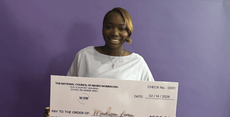 A student stands against a purple wall, holding a large ceremonial college scholarship check for $1,000 from the National Council of Negro Women DSU.