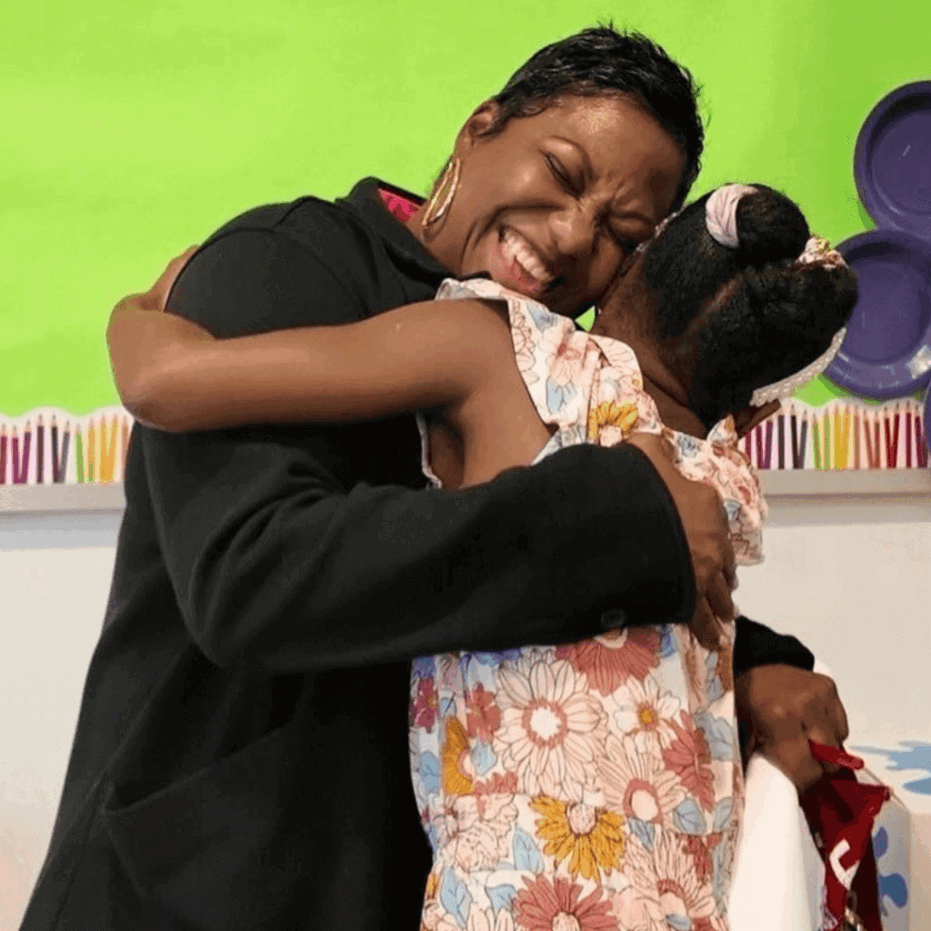An adult and a child share a joyful hug in a brightly decorated room, where the green bulletin board highlights student leadership and purple wall decorations add a lively backdrop.