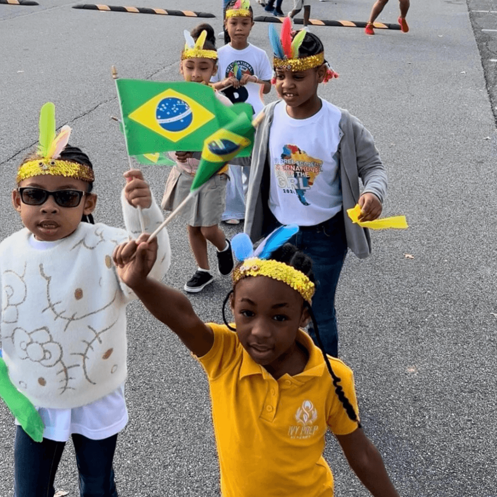 A group of children wearing feather headbands walk outdoors, showing leadership as one child proudly holds a Brazilian flag.