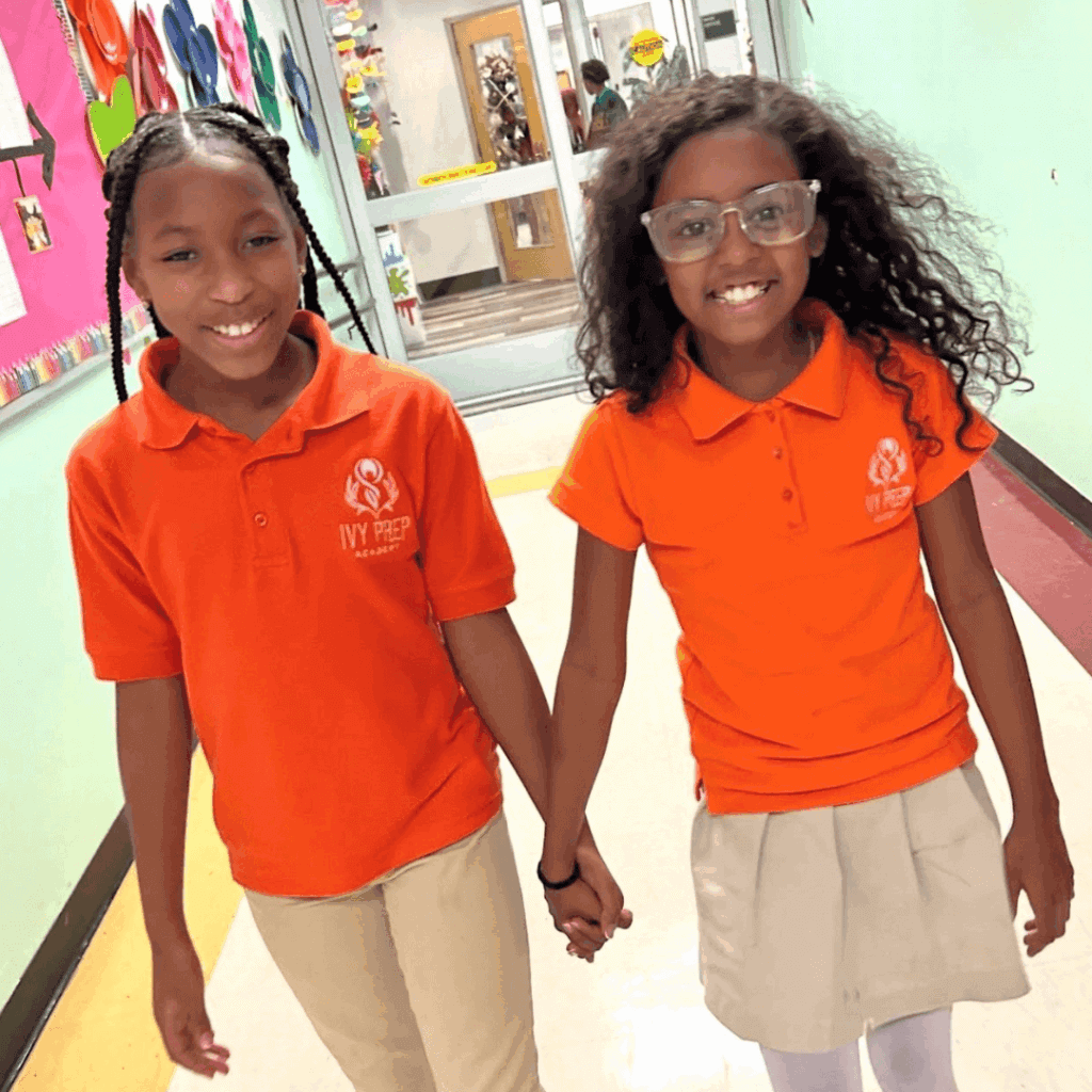 Two girls in orange Ivy Prep Academy uniforms hold hands and smile, showing leadership and friendship as they walk down a school hallway decorated with colorful artwork.