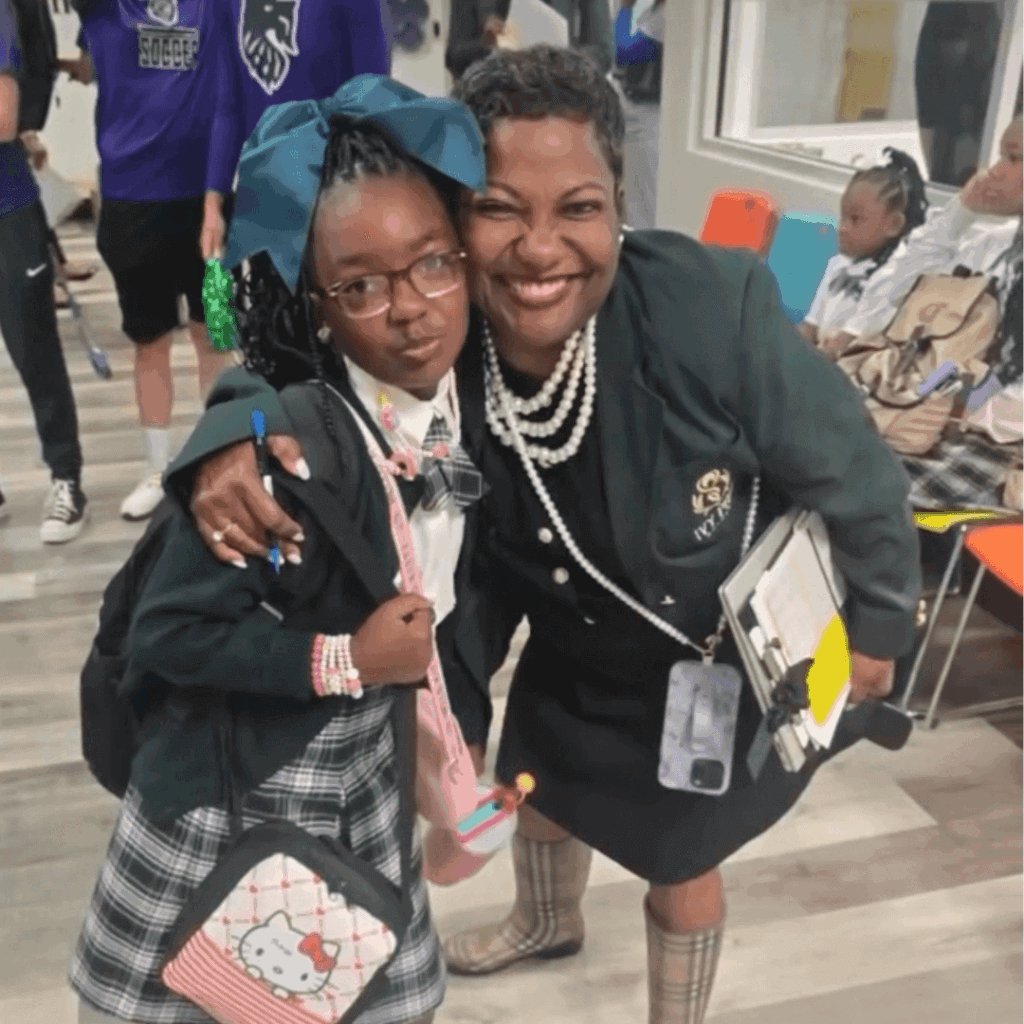 An adult and a child wearing school uniforms pose for a photo together indoors; displaying leadership, the smiling adult holds a clipboard while hugging the child.
