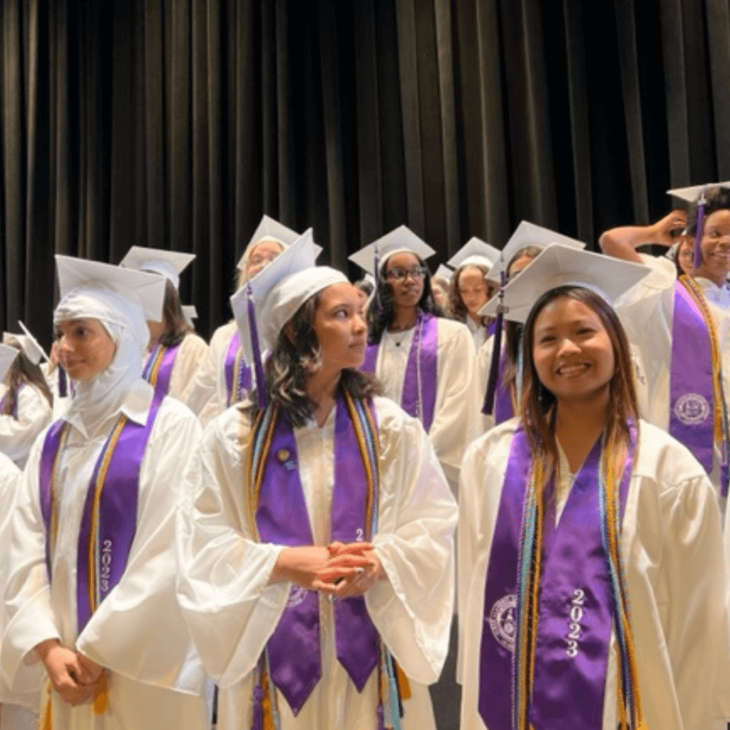 A group of students in white graduation gowns and purple stoles, some smiling, stand on stage during a graduation ceremony, celebrating their achievements and leadership.