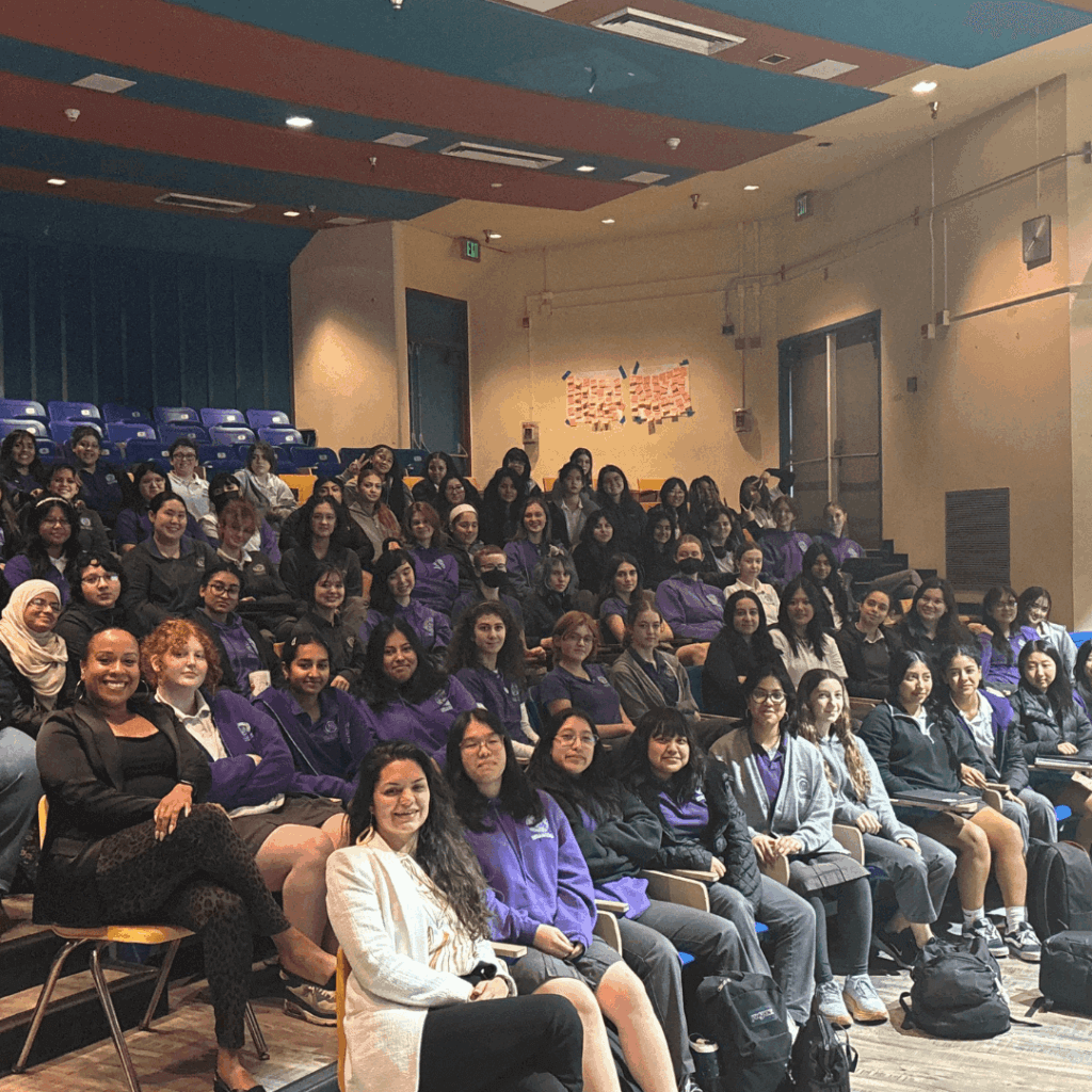 A large group of people, mostly women, sit in rows of chairs in an auditorium, posing for a group photo. Many are wearing purple tops or jackets, showcasing unity and leadership within the community.