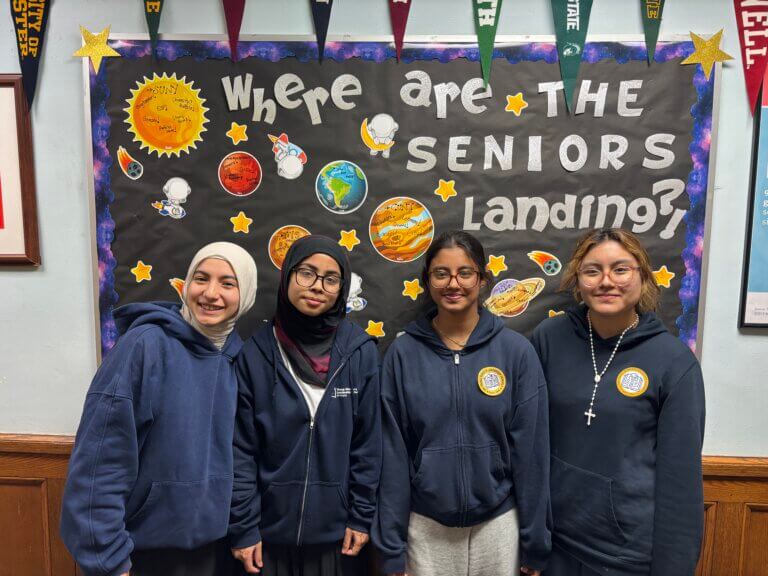 Four students in navy blue hoodies stand in front of a bulletin board decorated with planets and the text "Where are the seniors landing?" plus college pennants above, showcasing a student spotlight at TYWLS Astoria and how it’s producing stars.