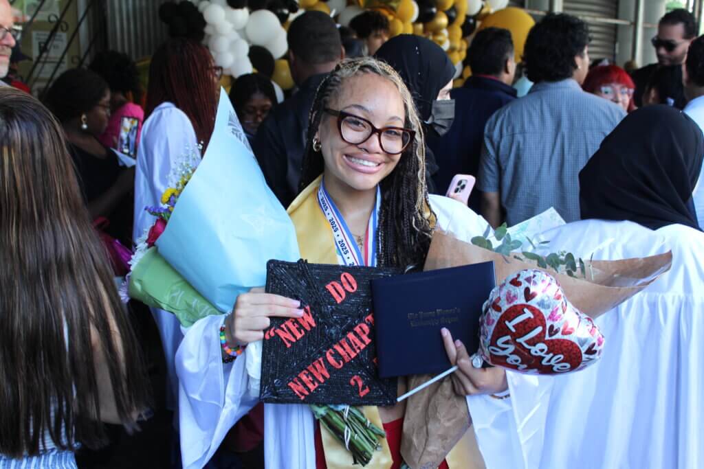 A young woman with braided hair and glasses is wearing graduation attire, holding a diploma, bouquet of flowers, decorated cap, and a heart-shaped balloon
