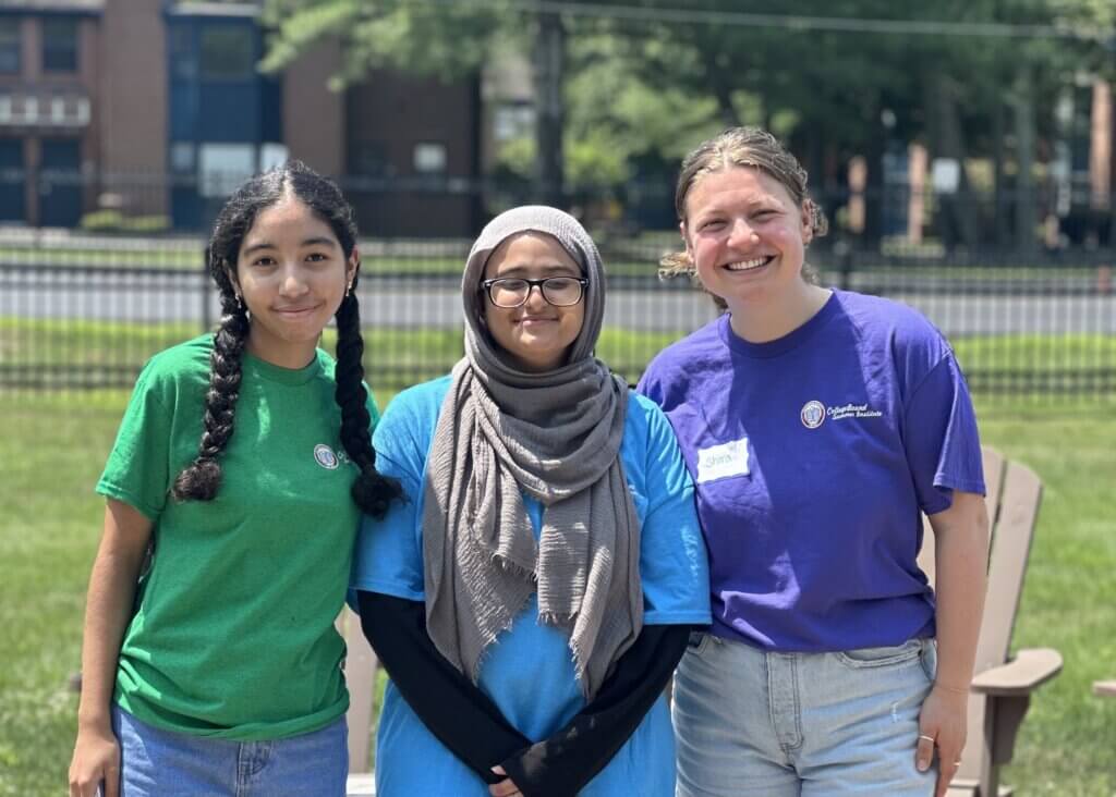 Three people stand outdoors on grass, smiling at the camera. Two wear colored t-shirts and one, a peer mentor, wears a hijab and glasses. A road and trees are visible in the background.