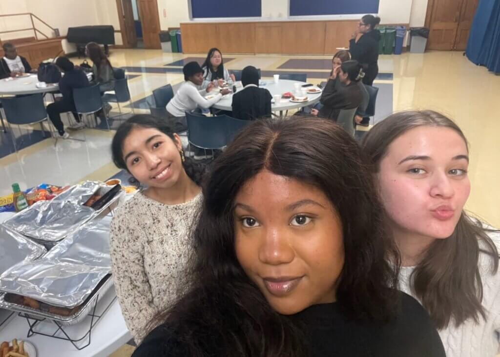 Three young women take a selfie in the foreground of a dining hall, with groups of people eating and talking at tables in the background.