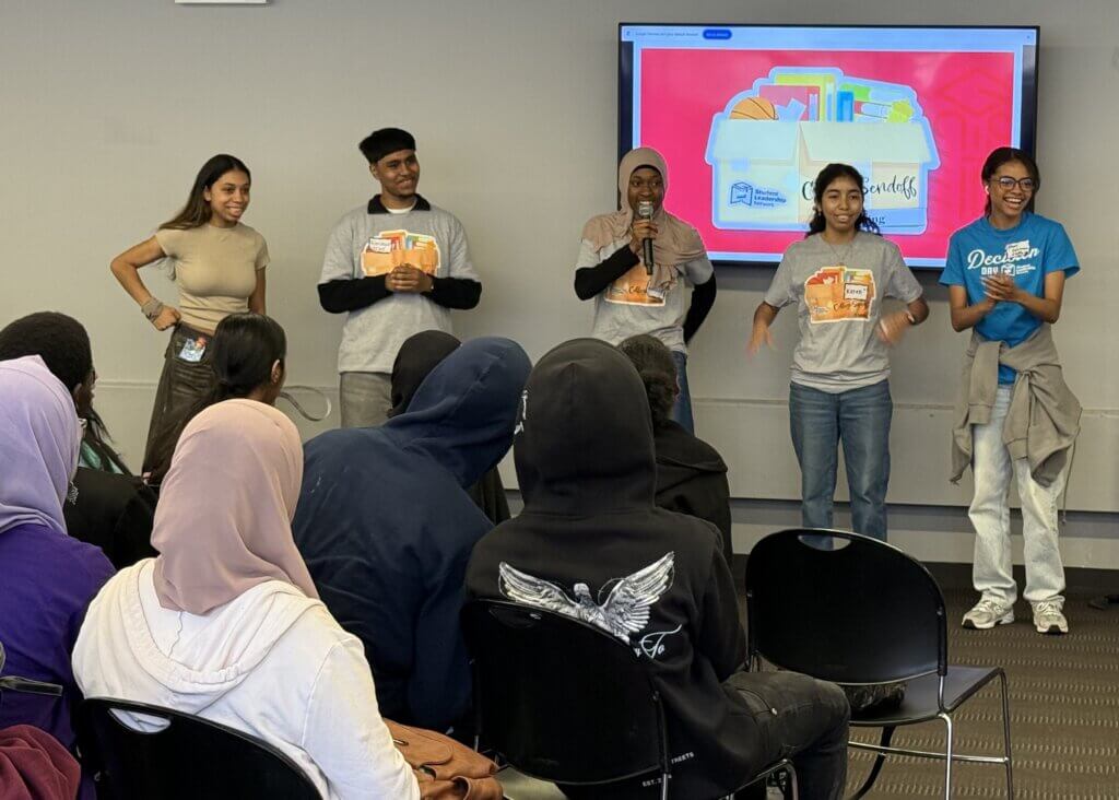 Five young peer mentors stand at the front of a room presenting to an audience, with a presentation slide displayed on a screen behind them.