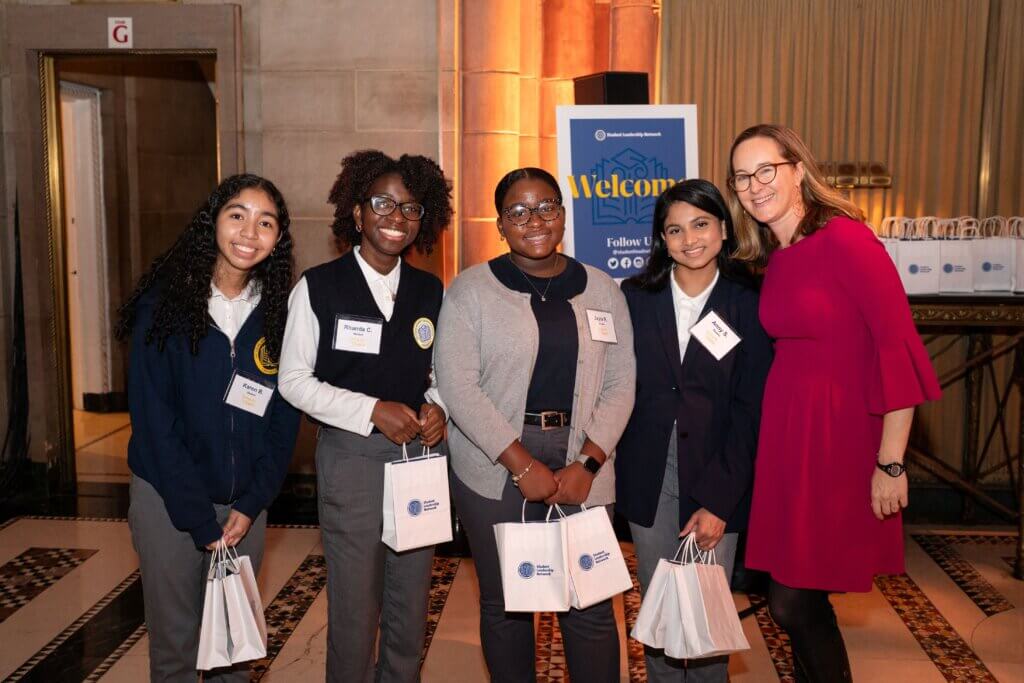 Five women posing indoors at an event, each holding a white gift bag and wearing name tags, with a “Welcome” sign and more bags visible in the background.