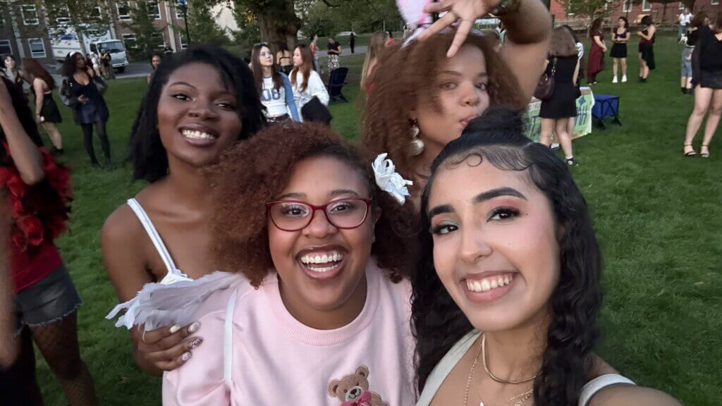 Four young women smile for a selfie at an outdoor gathering on a grassy area, with other people and trees visible in the background.