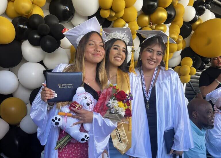 Three graduates in white gowns and caps smile at a ceremony, holding flowers, a diploma, a stuffed bear, and balloons in the background.