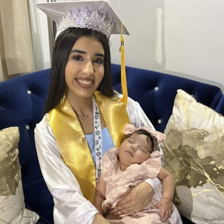A young woman in graduation attire and a tiara sits on a couch, smiling and holding her baby sister