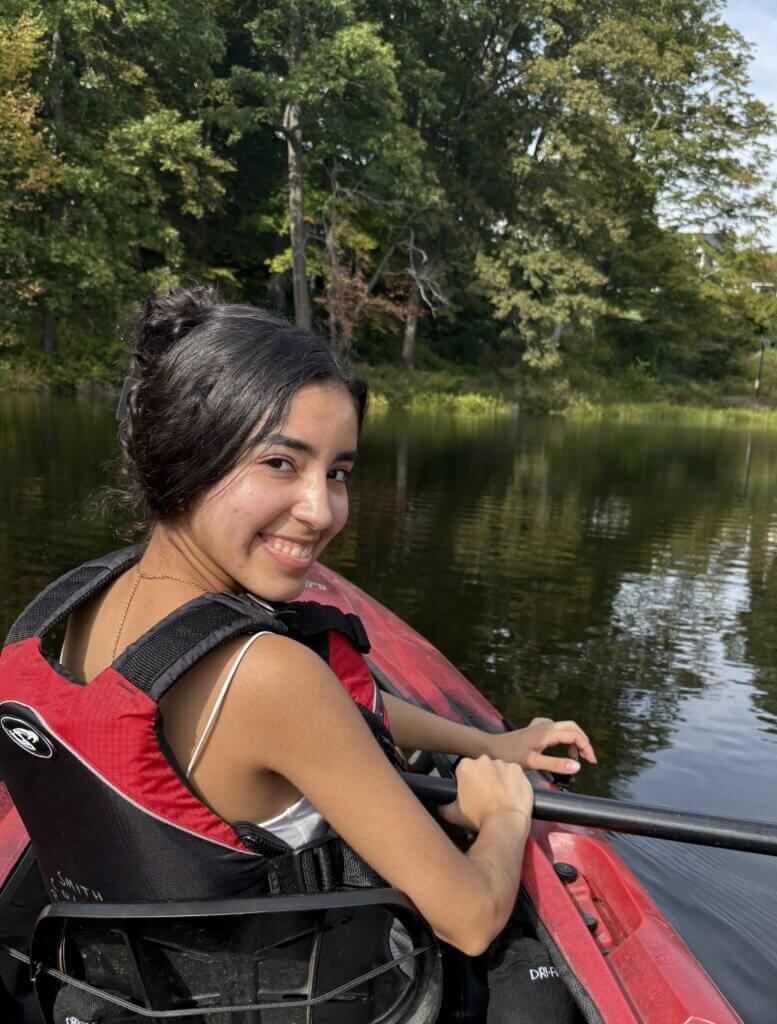 A person with dark hair in a red life jacket sits in a red kayak on calm water, smiling at the camera with trees in the background.