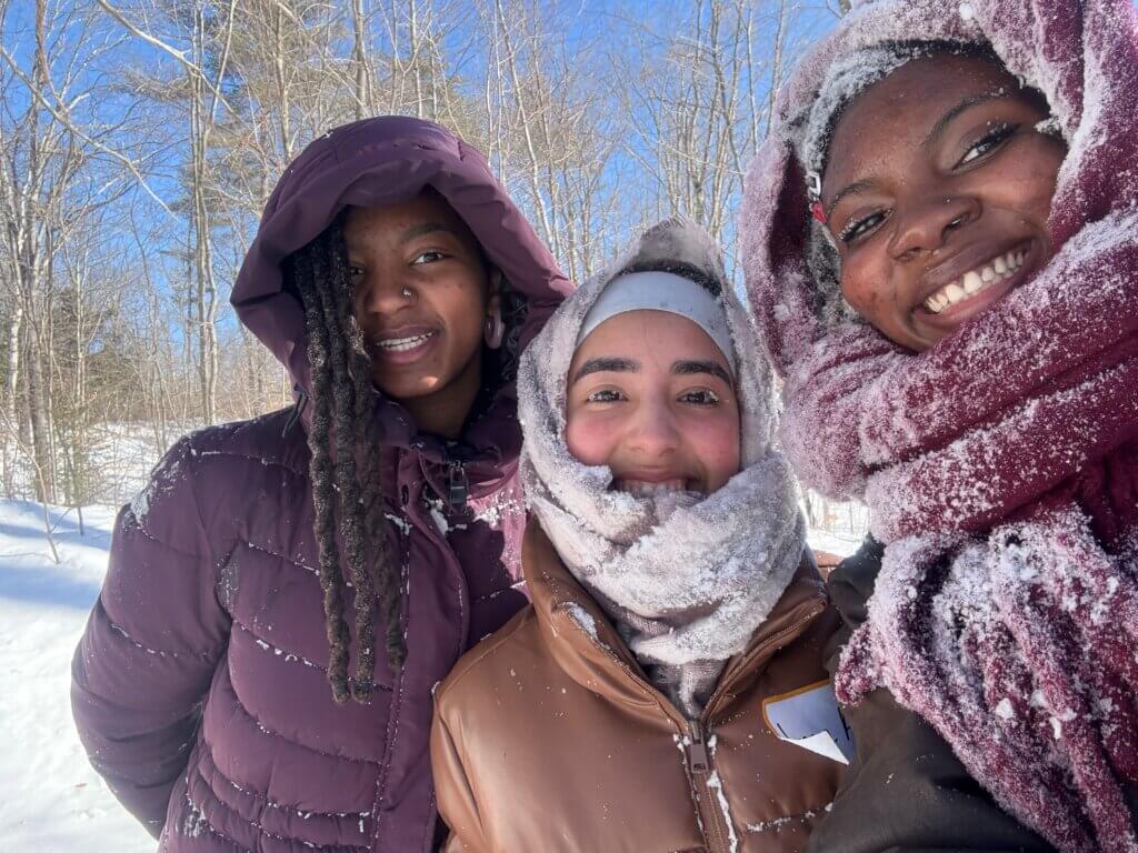 Three people bundled in winter clothing and scarves stand close together outdoors, smiling, with snow on their faces and jackets. Leafless trees and blue sky are visible in the background.