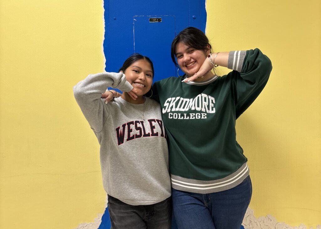 Two people stand smiling in front of a yellow and blue wall, wearing sweatshirts that say Wesley and Skidmore College.