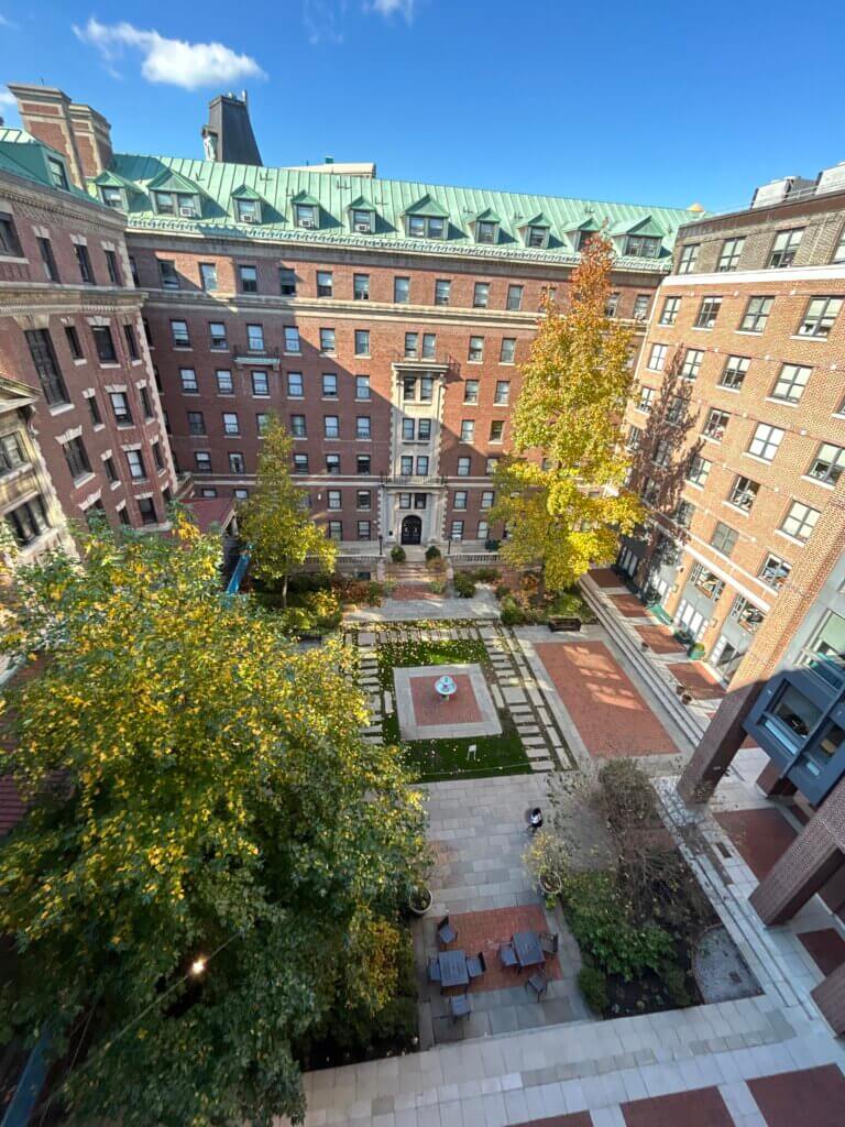 A view from a college dorm room window above of a rectangular courtyard with trees, pathways, a fountain in the center, and surrounding brick buildings under a clear blue sky.