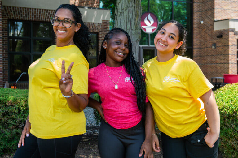 Three young women pose and smile outdoors in front of a college building during a summer program for first-gen students and other rising seniors in high school