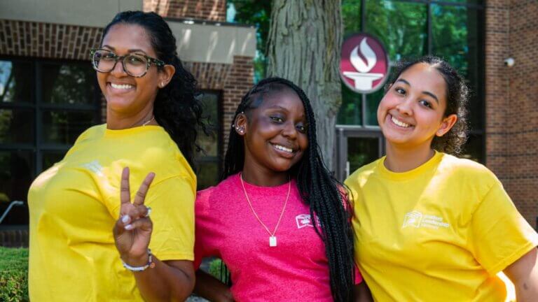 Three young women pose and smile outdoors in front of a college building during a summer program for first-gen students and other rising seniors in high school
