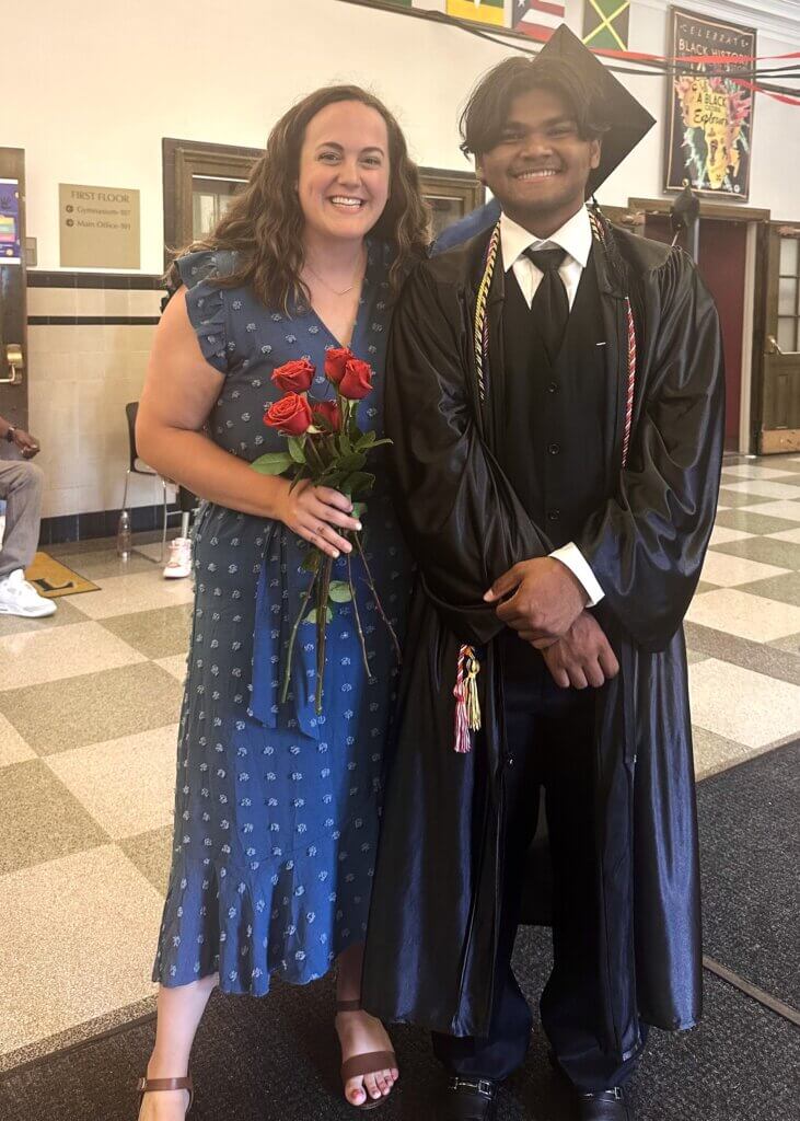 A college counselor holding roses stands next to a young man in a graduation cap and gown, both smiling, in a school hallway.