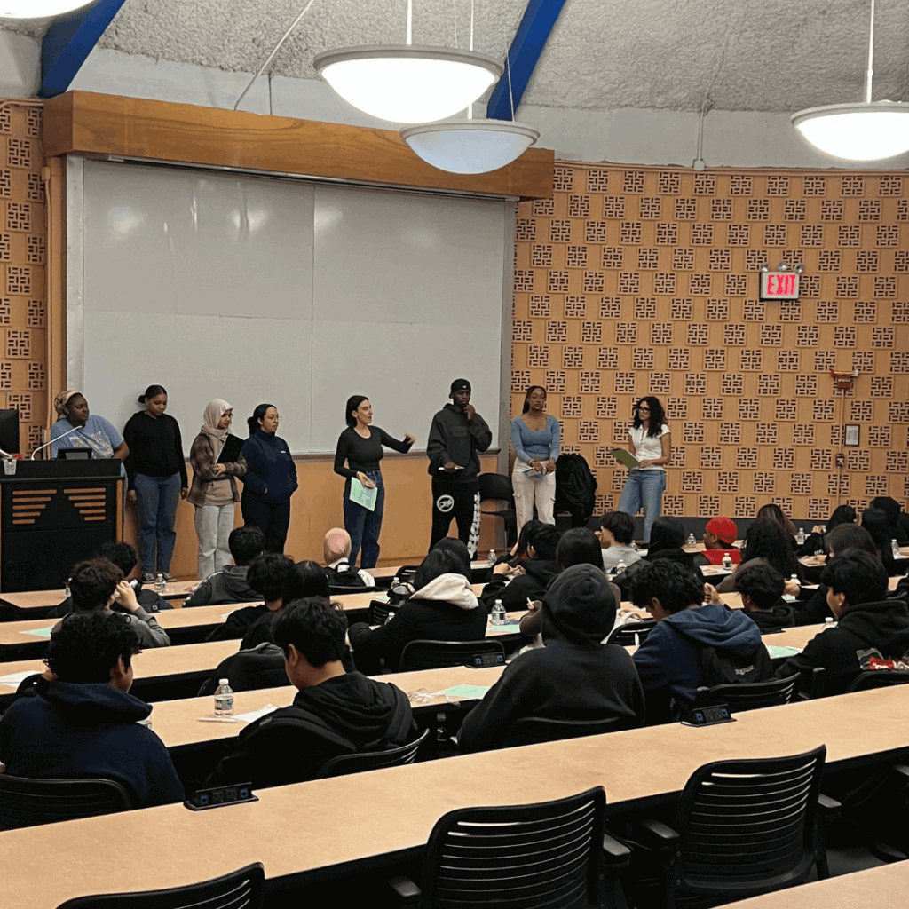 A group of people stands at the front of a lecture hall, demonstrating leadership as they face an audience seated at rows of desks. The academic setting features a whiteboard and podium.