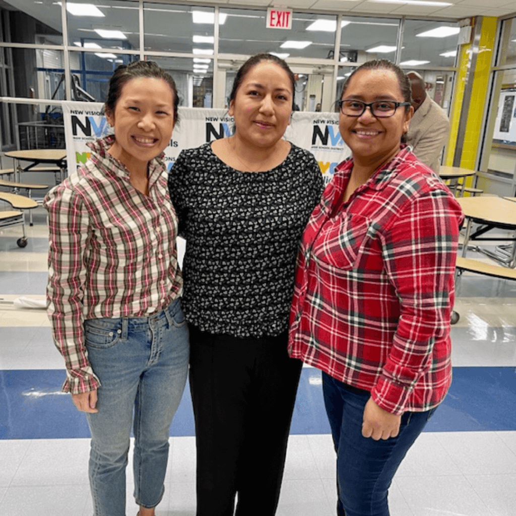 Three women, showcasing leadership, stand together in a cafeteria, smiling at the camera. Two wear plaid shirts, and one wears a black patterned top. Tables and windows can be seen in the background.