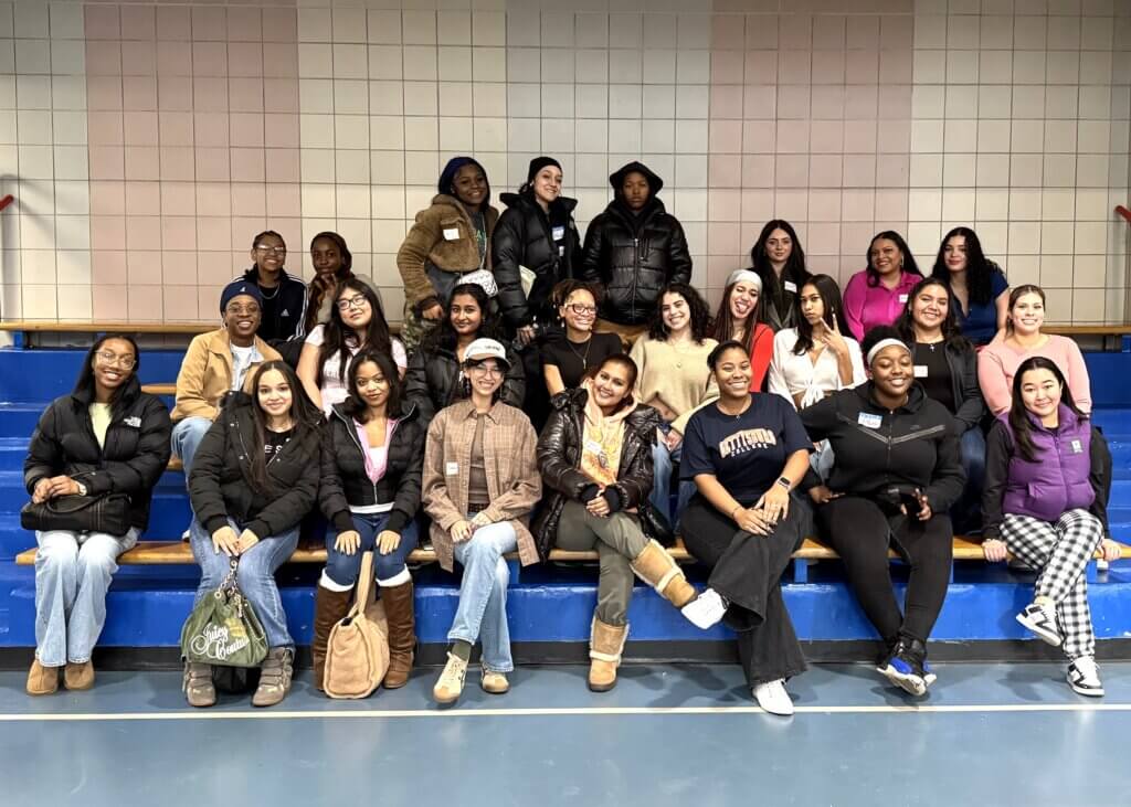 A group of TYWLS teachers, alumni, and staff sit and stand on wooden bleachers in a gymnasium, posing for a group photo during Alumni Day