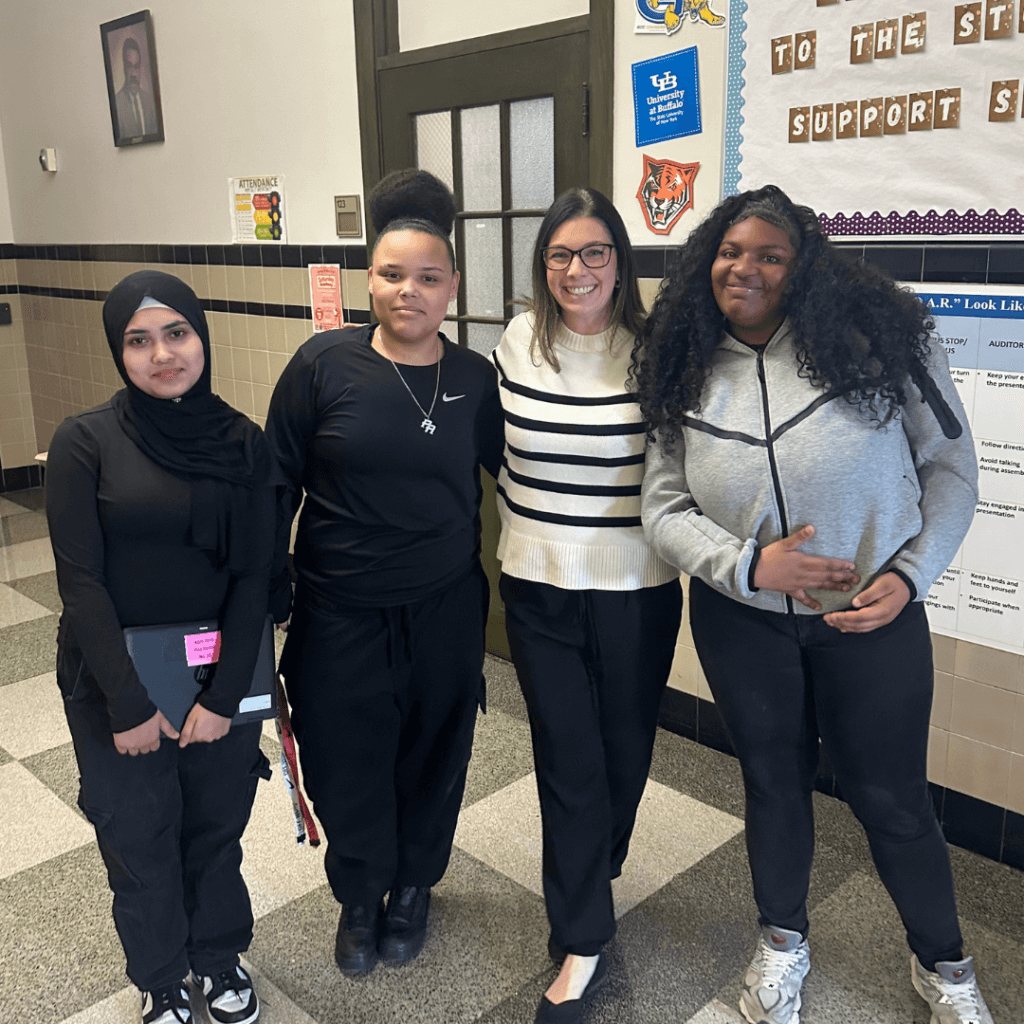 Four women stand together in a school hallway, showcasing their leadership as they pose for a group photo in front of bulletin boards and closed doors.