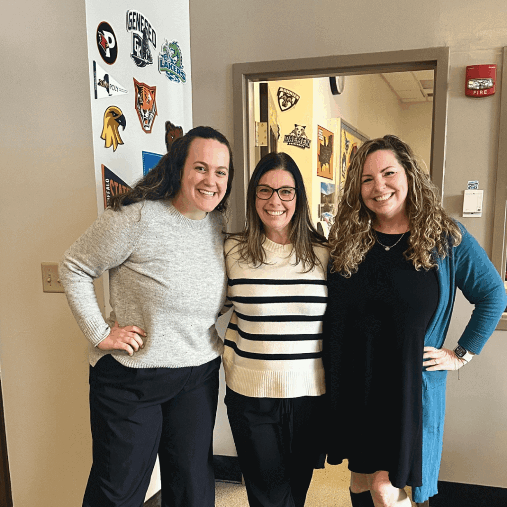 Three women stand side by side smiling indoors, near a wall decorated with various school or team logos, showcasing their leadership and camaraderie.