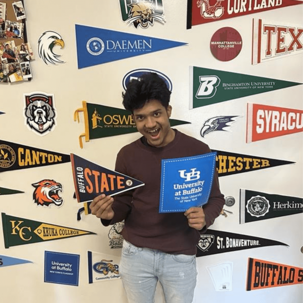 A person stands in front of a wall decorated with college pennants, holding Buffalo State and University at Buffalo pennants, smiling at the camera and proudly showcasing their leadership aspirations.