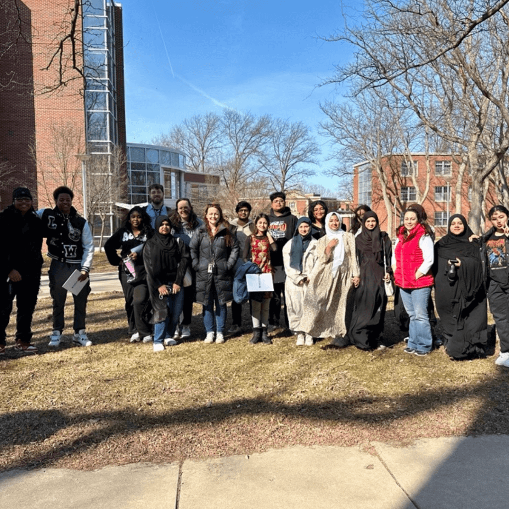 A group of people, united by their leadership, pose for a photo outdoors on a sunny day, with campus buildings and bare trees visible in the background.