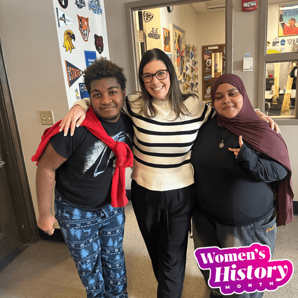 Three people stand together and smile in a school hallway decorated with posters, highlighting leadership and unity. A Women’s History Month graphic appears in the lower right corner.