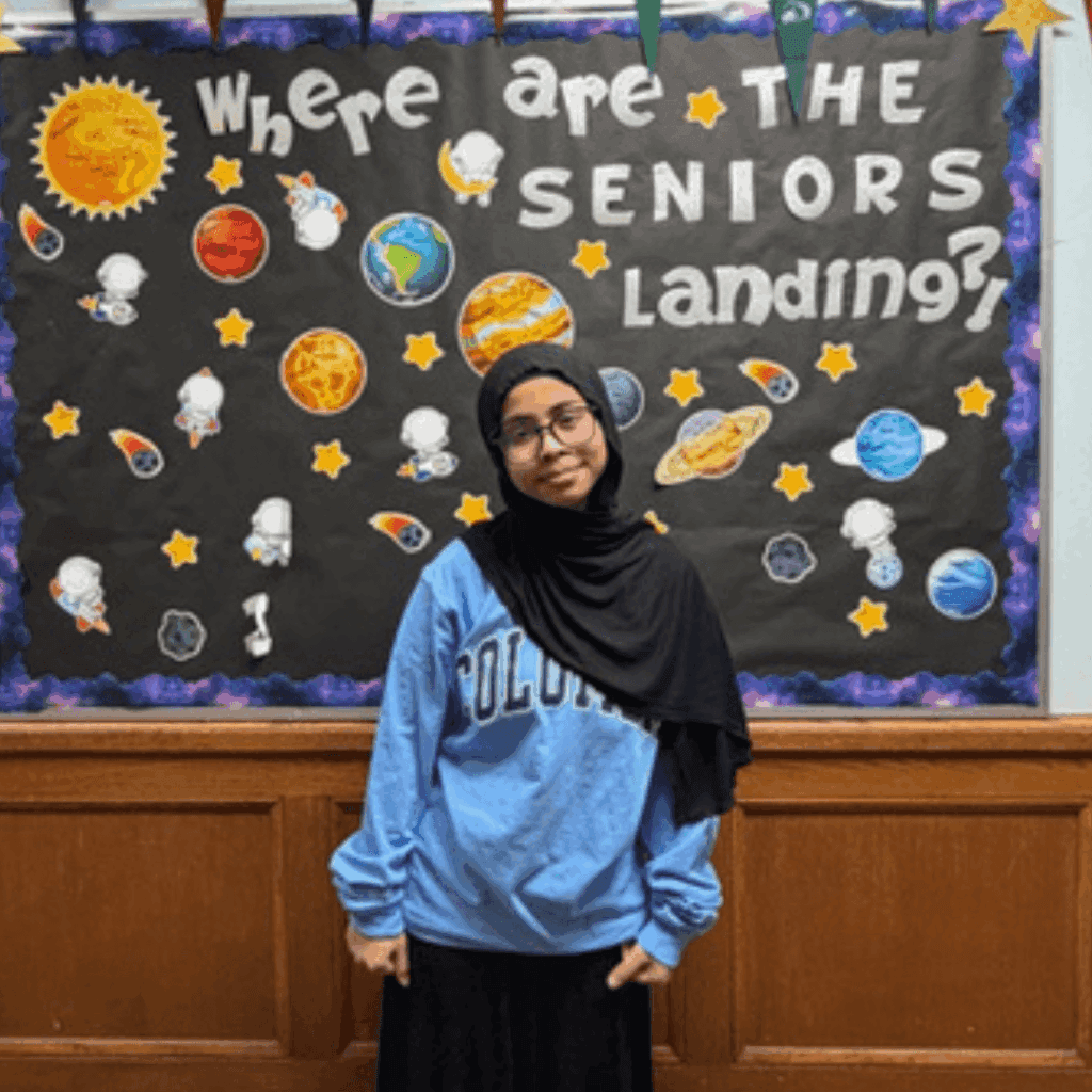 A student in a light blue Columbia sweatshirt stands in front of a TYWLS Astoria bulletin board that reads Where are the seniors landing? with space-themed decorations, producing stars in this student spotlight moment.