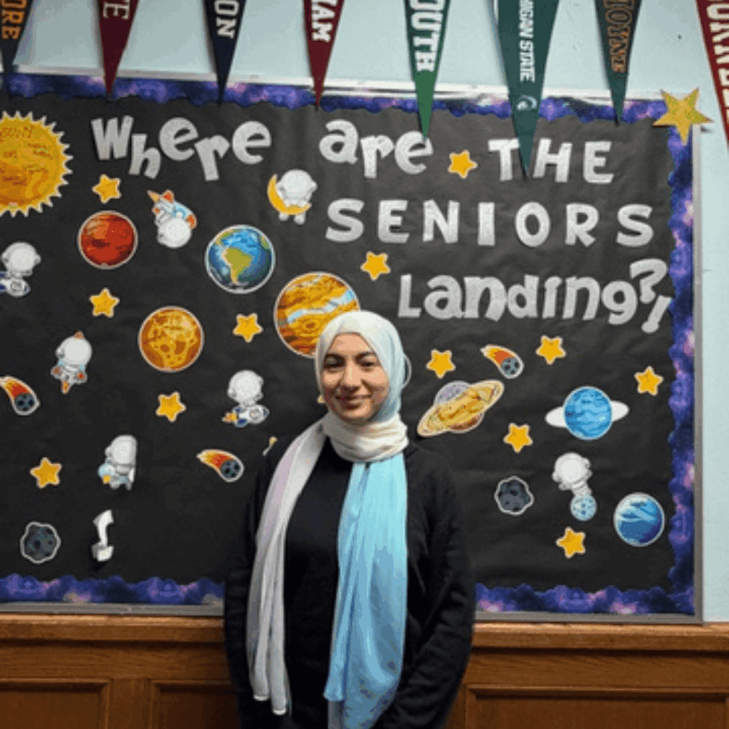 A person in a light blue and white hijab stands in front of a TYWLS Astoria bulletin board that says Where are the seniors landing? decorated with planets and college pennants, highlighting the student spotlight and producing stars of the future.