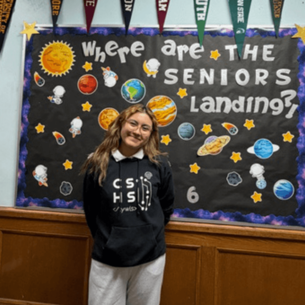 A Student Spotlight shines as a student stands before the TYWLS Astoria bulletin board that reads "Where are the seniors landing?" surrounded by planets and space-themed decorations, truly producing stars for the future.