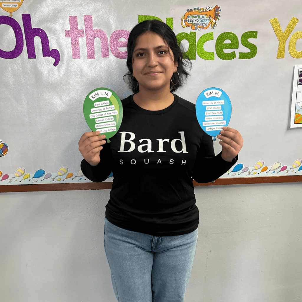 A person in a Bard Squash shirt stands in front of a decorated wall, holding two colored paper cutouts with text, embodying college dreams and the spirit of achievement.