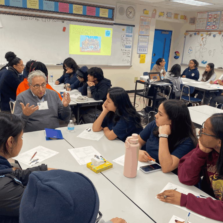 A group of students sits around tables listening to Jeffrey Gurual speak during a classroom discussion, with presentation slides and posters about education and opportunity visible in the background.