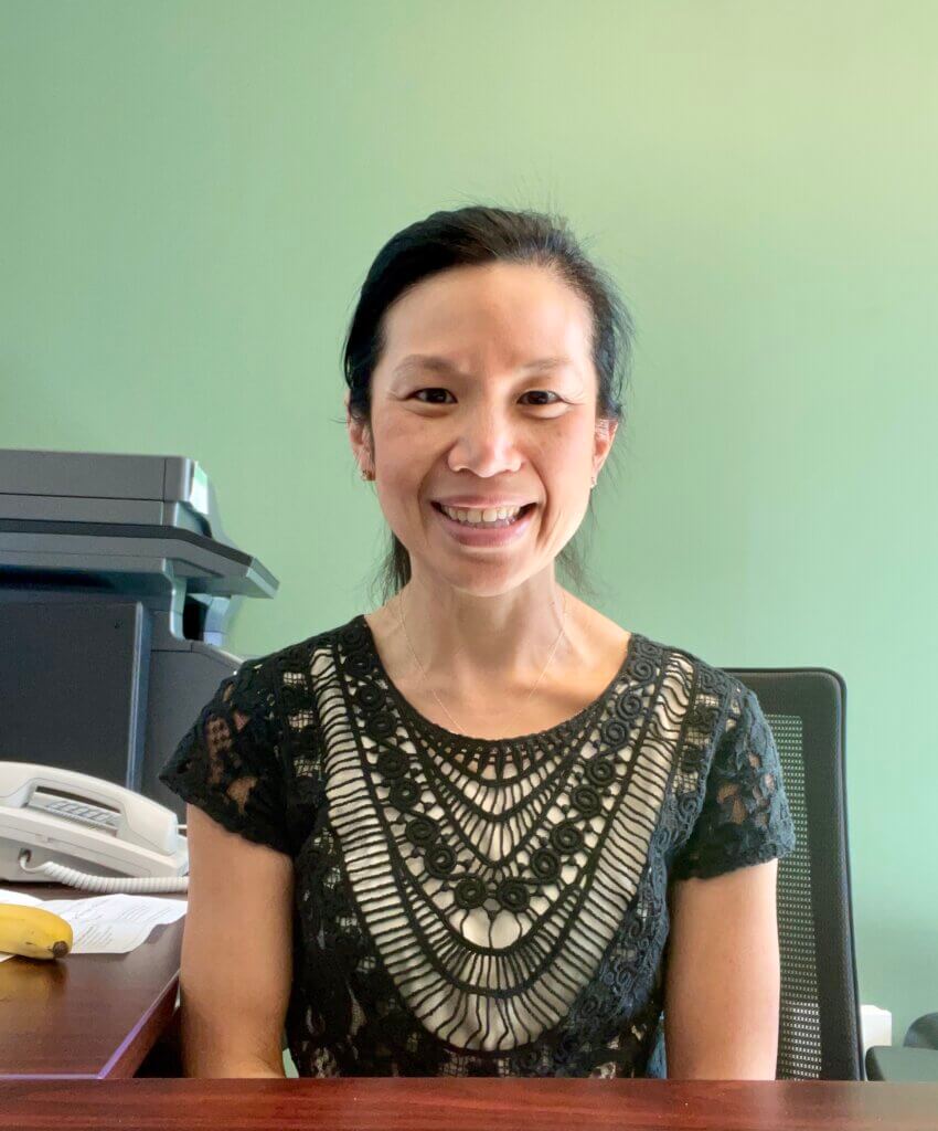A woman with dark hair tied back, wearing a black and white patterned top, sits at an office desk with a phone, printer, and a banana in the background, embodying quiet leadership in her professional workspace.