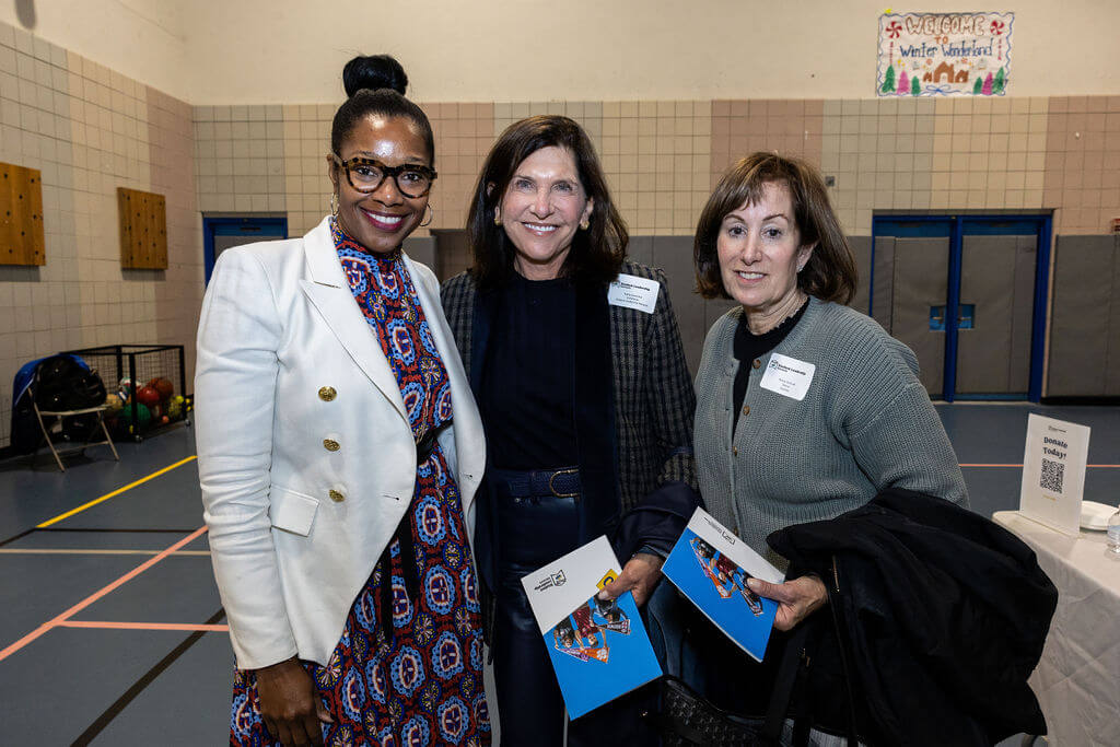 Three women stand together in a gymnasium, each holding a program booklet and wearing name tags, posing for a photo at an indoor event.