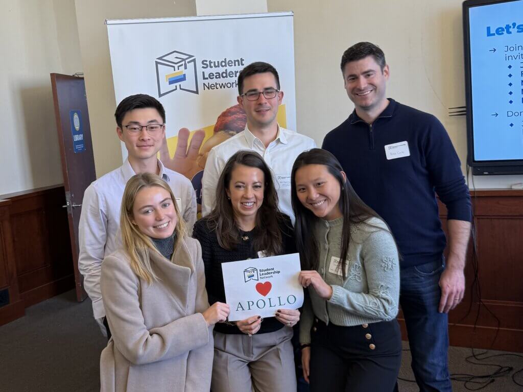 Six people pose indoors at a Student Leadership Network event, with three holding a sign that reads I ♥ APOLLO.
